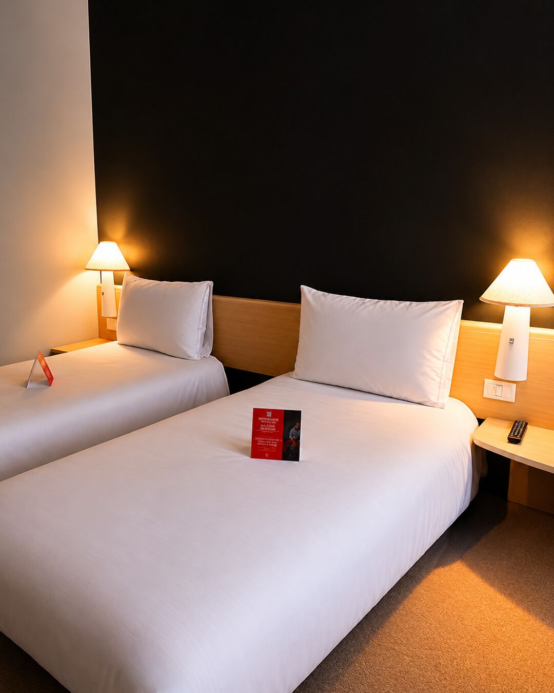 Habitación de hotel con dos camas, cada una con ropa blanca y almohadas, una mesita de noche con lámpara y una tarjeta informativa roja sobre la cama.
