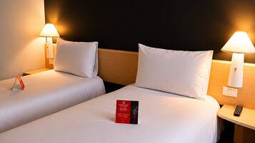 Habitación de hotel con dos camas, cada una con ropa blanca y almohadas, una mesita de noche con lámpara y una tarjeta informativa roja sobre la cama.