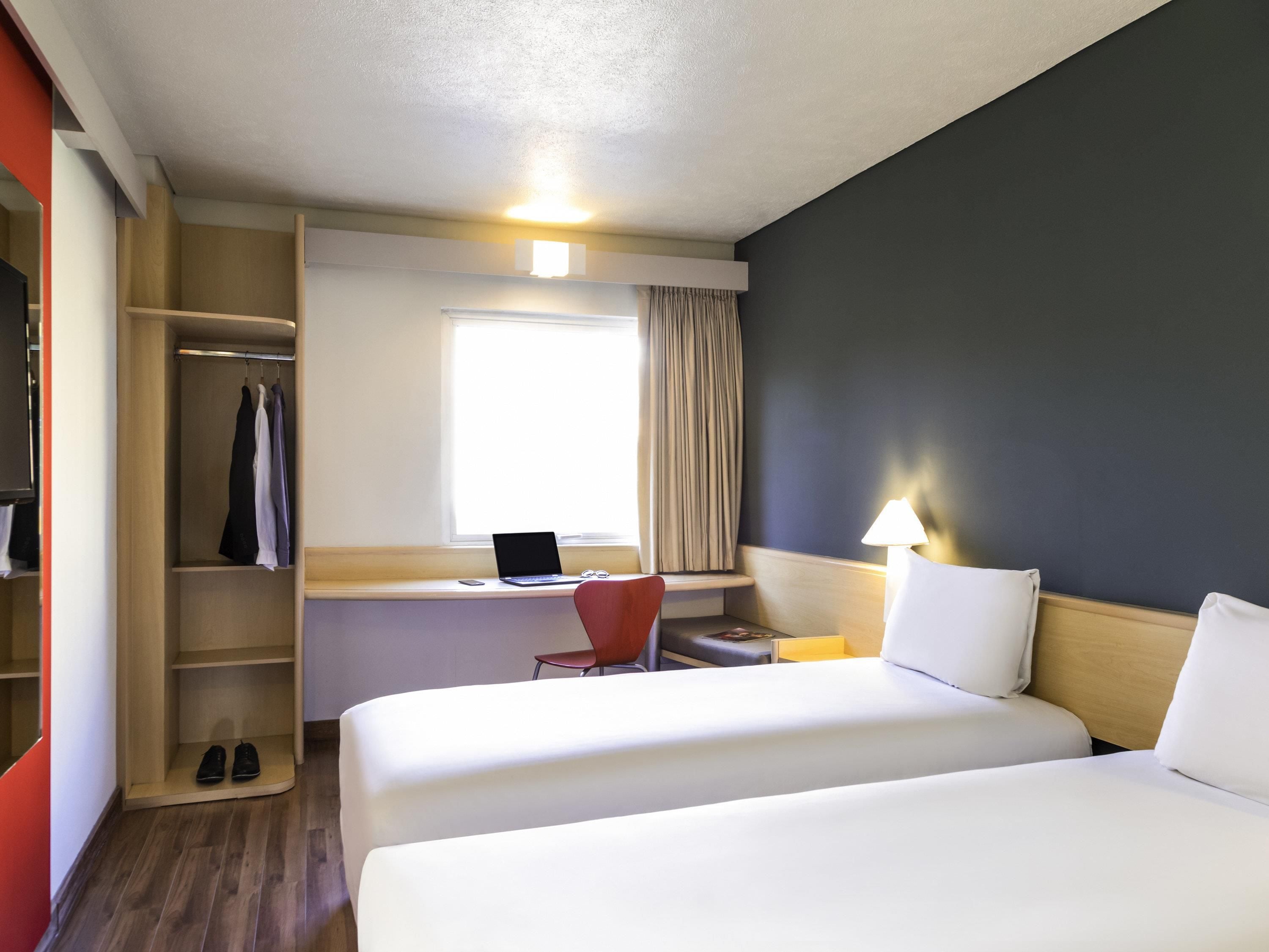 Habitación de hotel con dos camas, un escritorio con una computadora portátil, una silla, un armario y una ventana con cortinas.