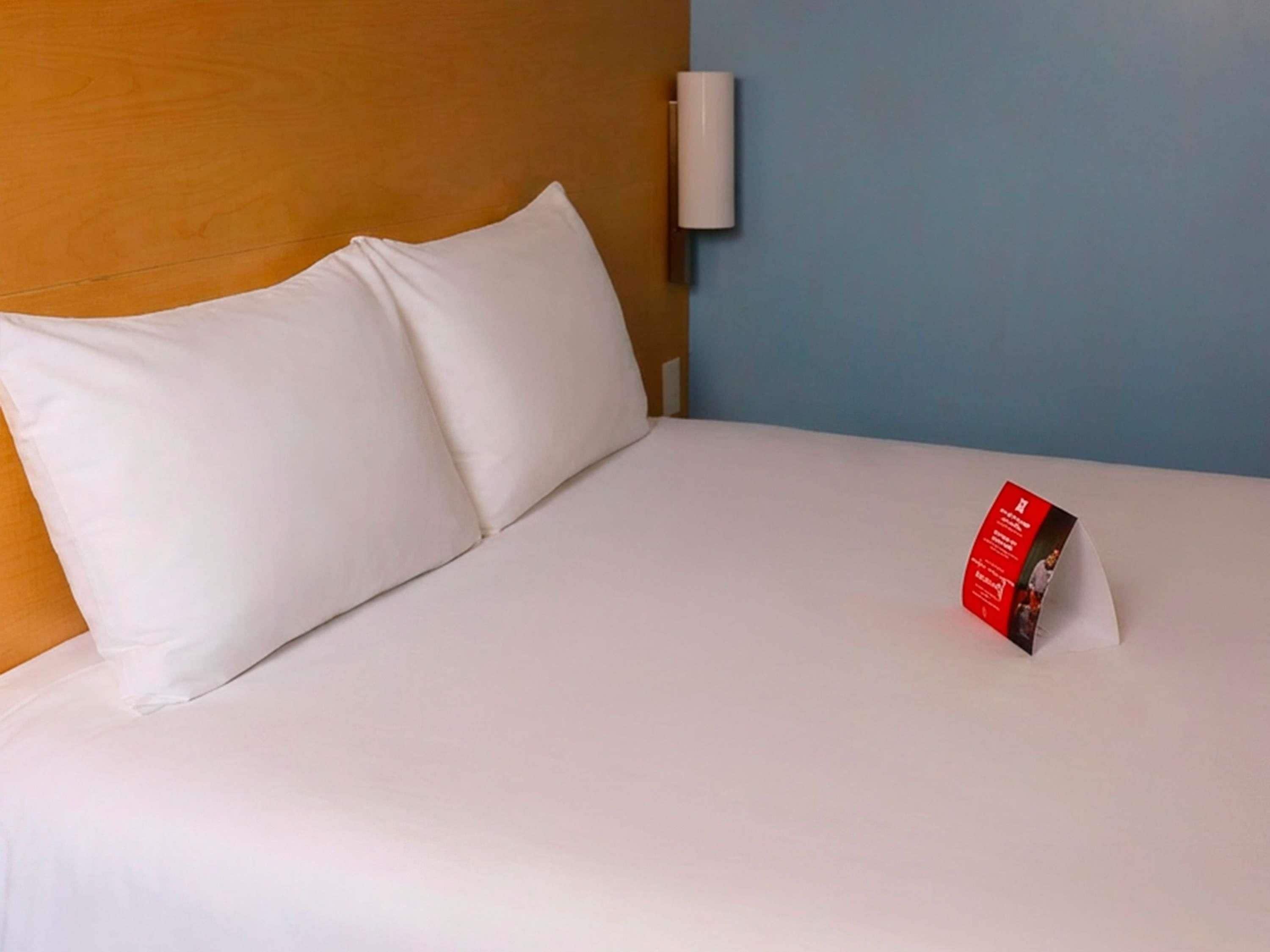 Een bed met witte lakens, een rood boek aan de rechterkant en een wandlamp.