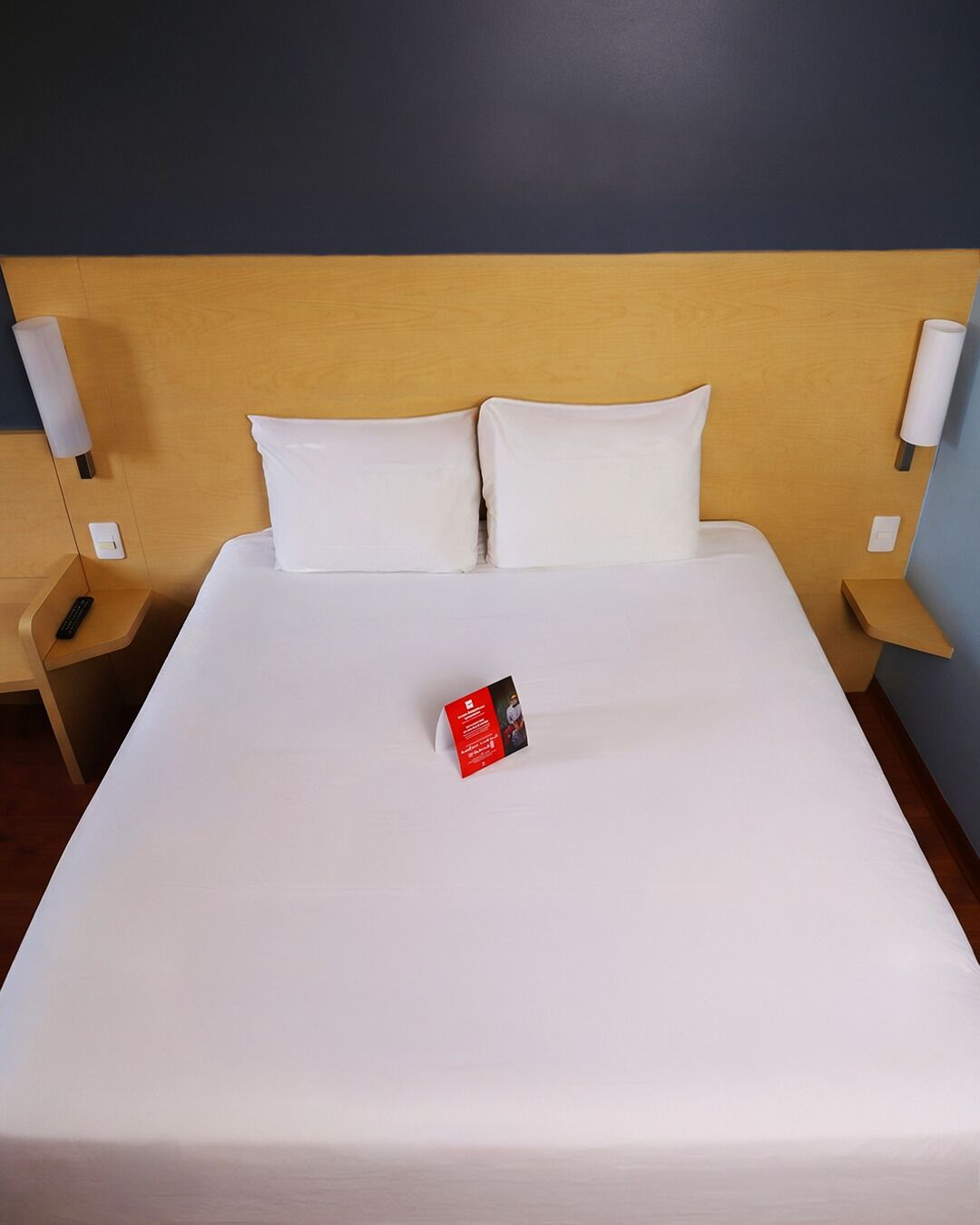 A neatly made bed with white linens, a remote control on the left side, and a brochure on the bed.