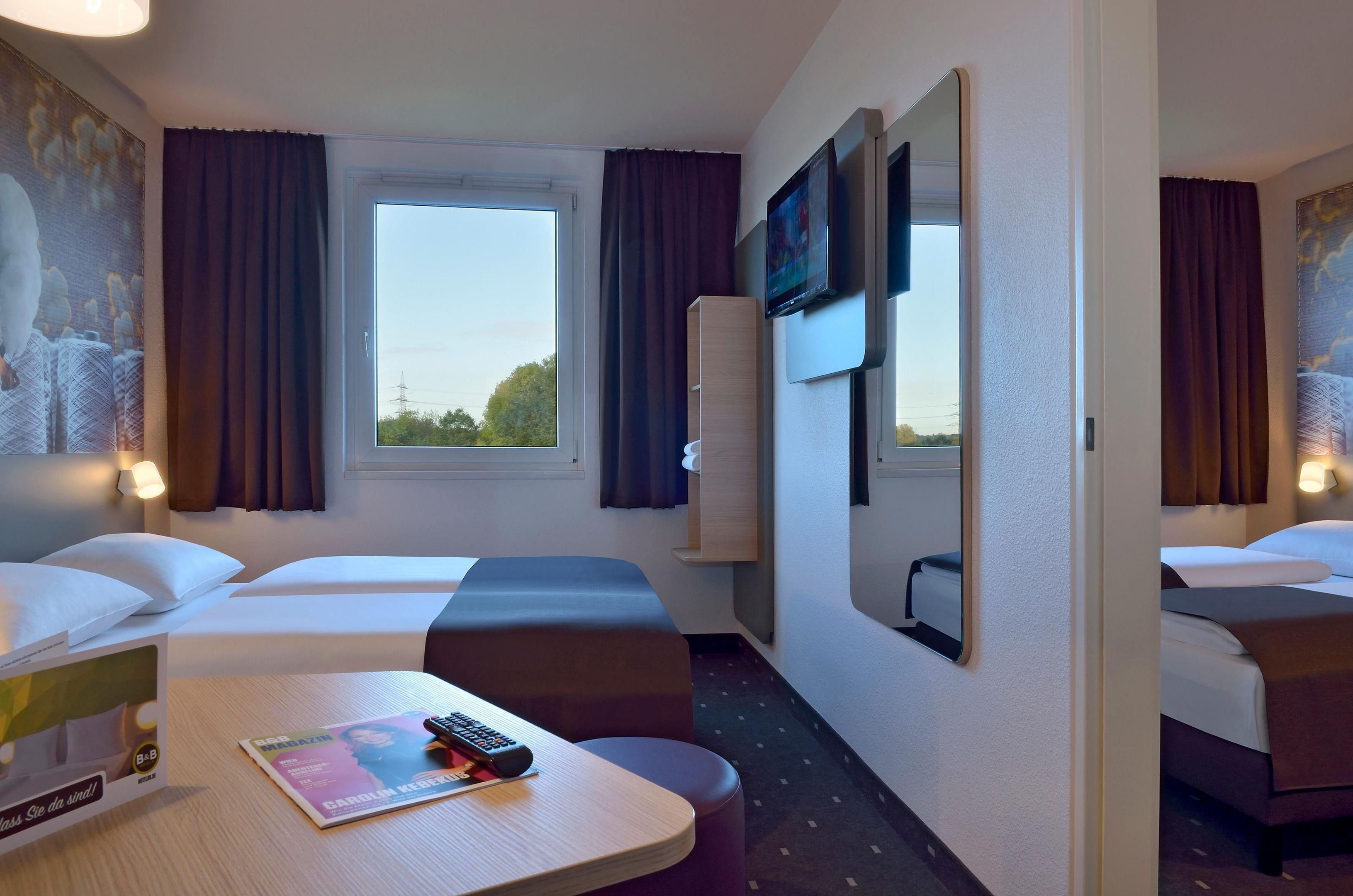 A hotel room with two beds, a window, a mirror, a table with a magazine and remote control, and a wall-mounted TV.