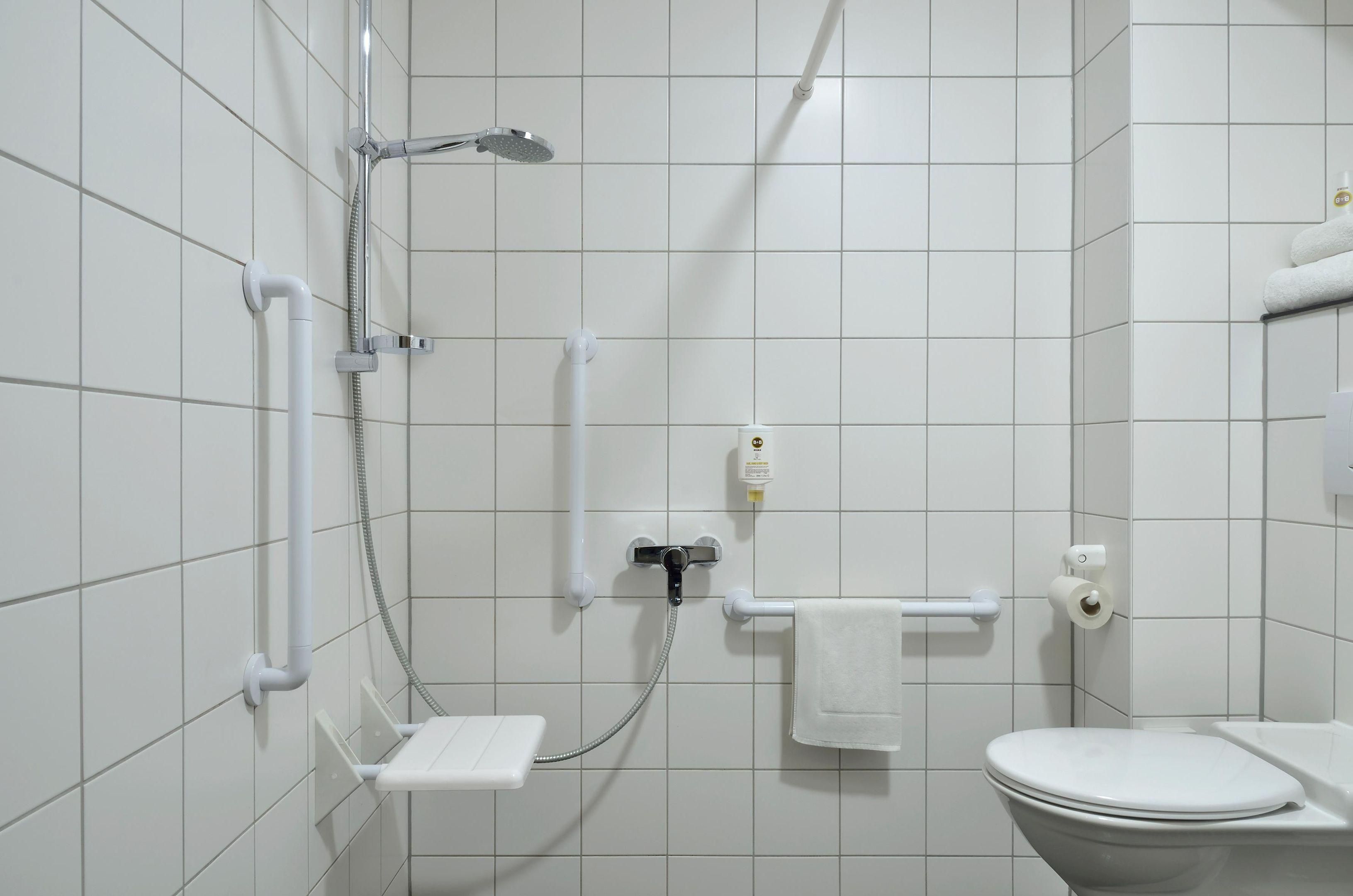 A bathroom with white tiles, a toilet, a shower with a handheld showerhead, a towel rack, and a soap dispenser.