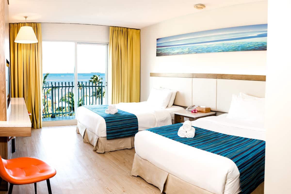 A hotel room with two beds, a desk, and a chair. There is a view of the ocean through the window.