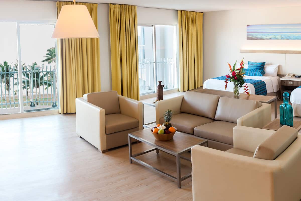 A hotel room with a sofa, armchairs, a coffee table with fruit, a bed with blue bedding, a nightstand, and a painting on the wall.