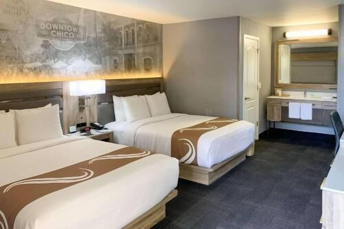 A hotel room with two beds, a nightstand, a lamp, a mirror, and a bathroom with a sink and towel rack.