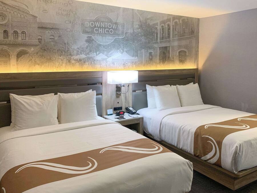 A hotel room with two beds, a nightstand with a lamp, and a wall mural depicting downtown Chico.
