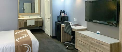 Habitación de hotel con cama, escritorio, televisor, espejo y baño con lavabo y toalla.