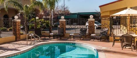 A swimming pool with lounge chairs and umbrellas, surrounded by palm trees and a gated fence.