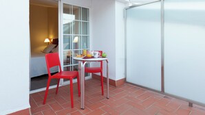 Ein kleiner Balkon mit zwei roten Stühlen, einem Tisch mit Frühstücksgebäck und eine Glasschiebetür, die in ein Schlafzimmer führt.