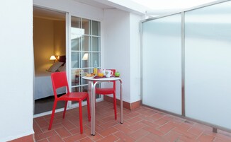 Un pequeño balcón con dos sillas rojas, una mesa con provisiones para el desayuno y una puerta corrediza de vidrio que da a un dormitorio.