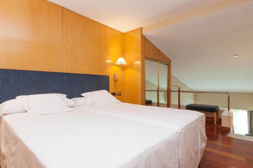 A modern hotel room with a large bed, wooden panel walls, a nightstand, a lamp, and a view of a balcony.