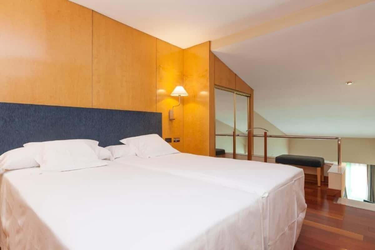 Una habitación de hotel moderna con una cama grande, paneles de madera, una mesita de noche, una lámpara y vistas al exterior.