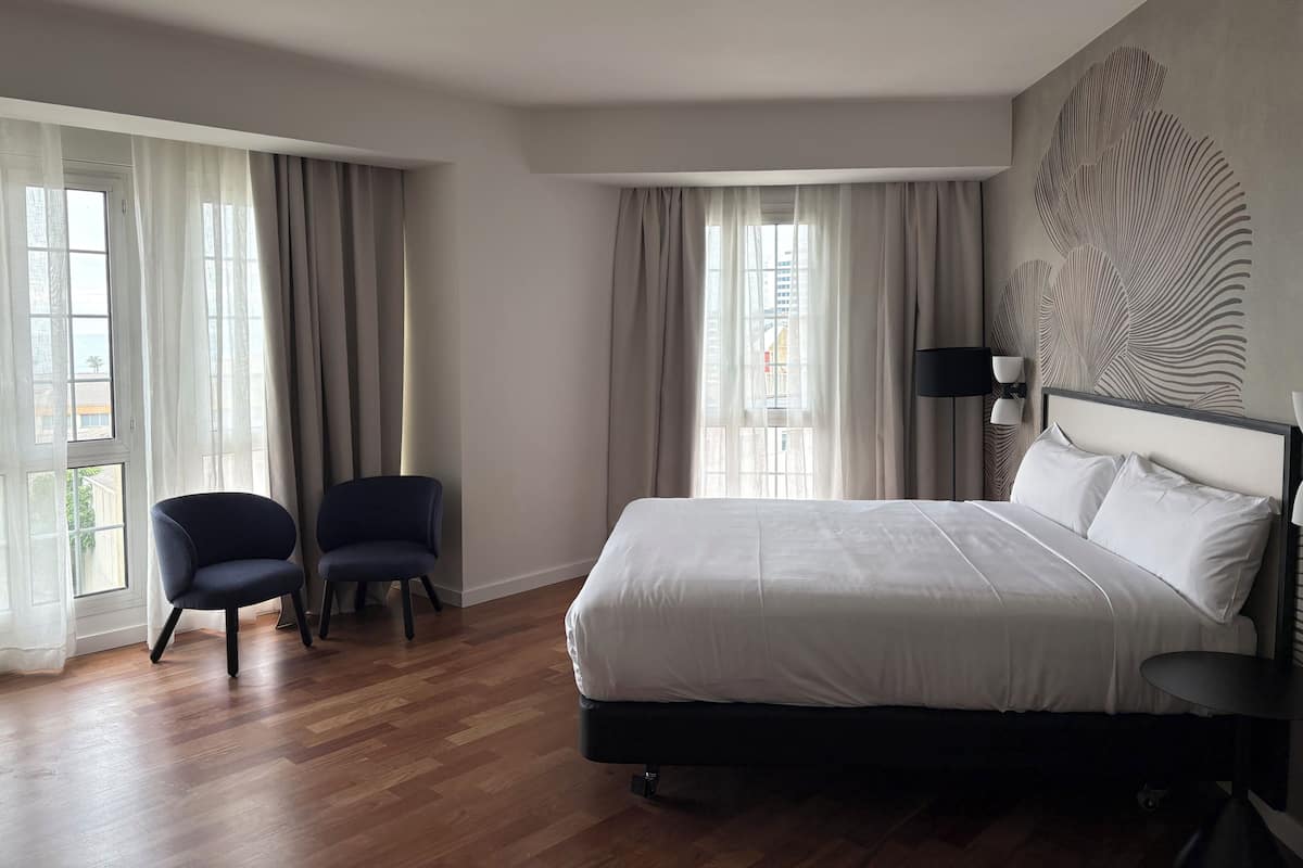 Habitación de hotel con una cama grande, dos sillones, un escritorio y vistas de la ciudad a través de las ventanas.