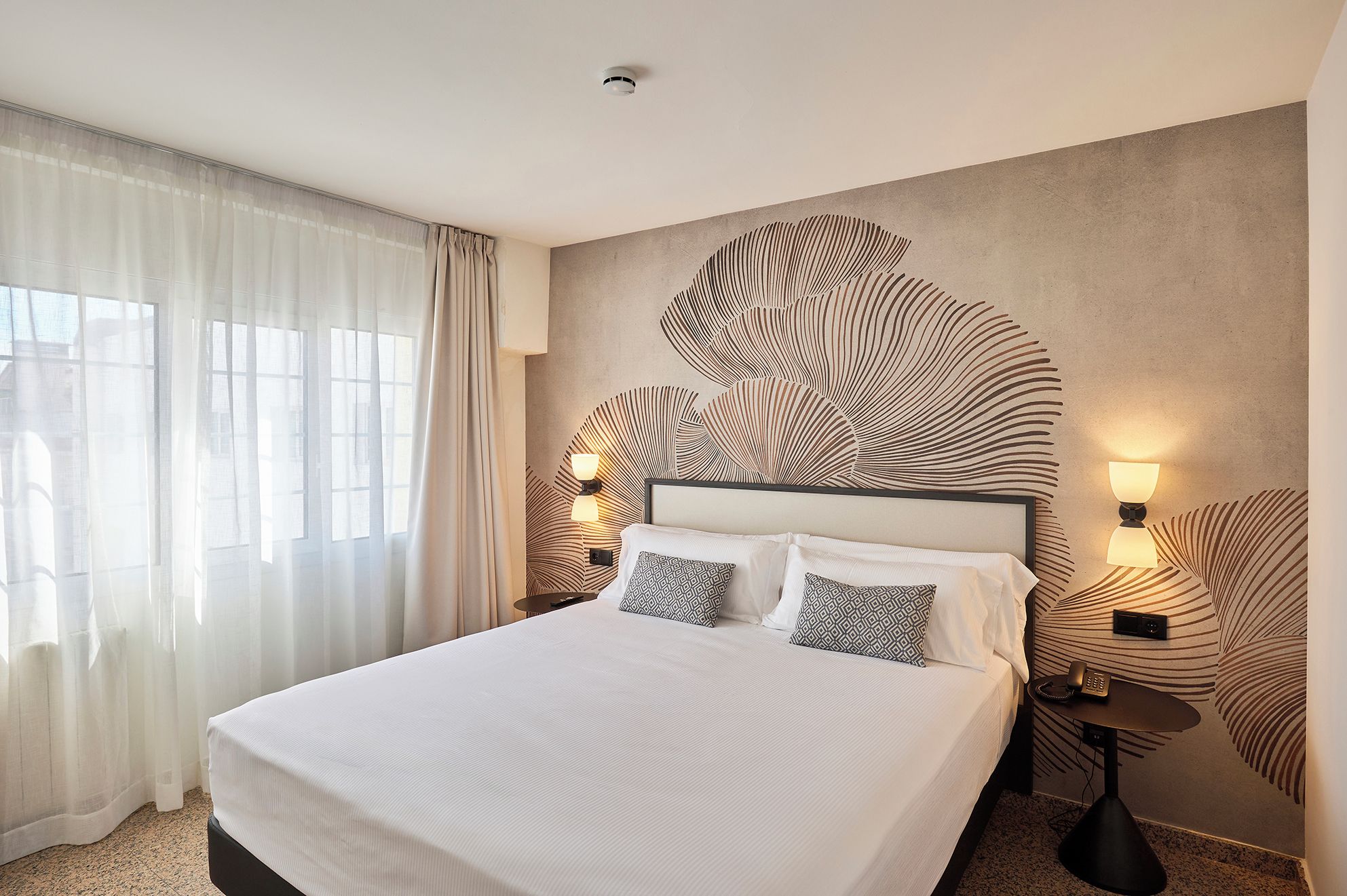 A hotel room with a large bed, two bedside lamps, a wall-mounted phone, and a window with sheer curtains.