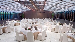 Une salle de banquet spacieuse, dotée de tables rondes et de chaises blanches, avec un plafond haut orné de guirlandes lumineuses et un mur incurvé agrémenté de fenêtres.