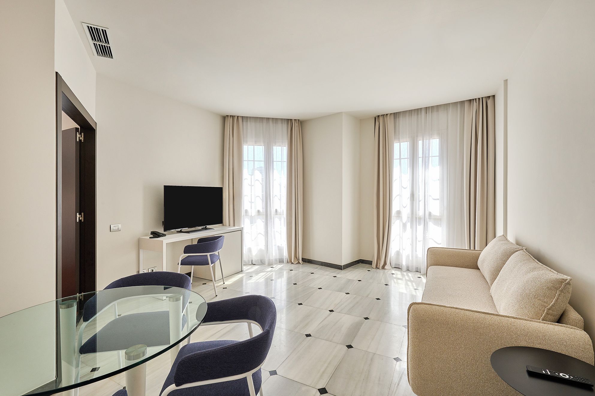 Una habitación de hotel moderna con una mesa de comedor de cristal, un sofá, un televisor y un escritorio con una silla.