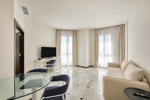 Une chambre d’hôtel moderne avec une table à manger en verre, un canapé, une télévision et un bureau avec une chaise.