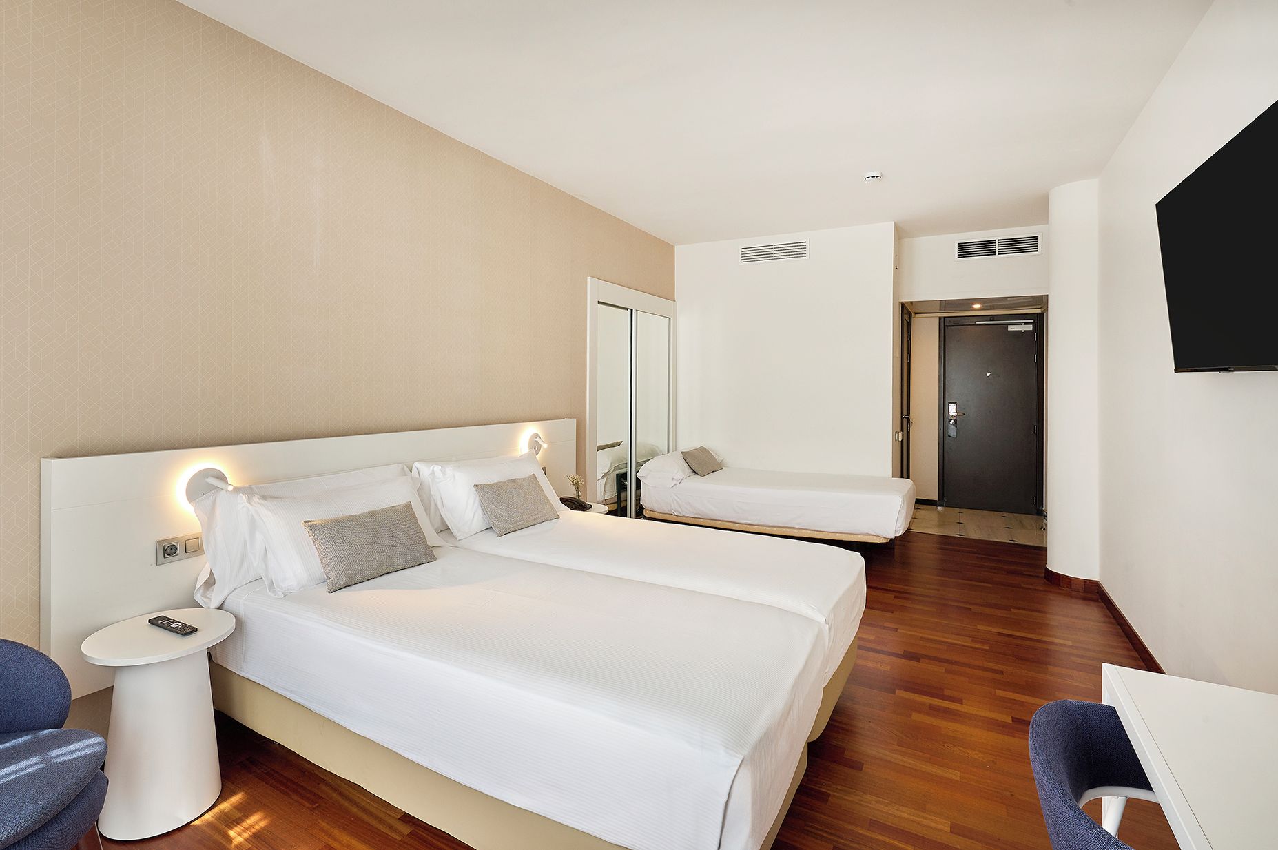 Quarto de hotel com duas camas, uma escrivaninha, uma cadeira, uma televisão e um guarda-roupa.