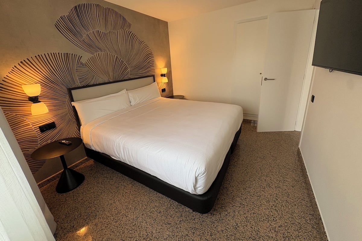 Una cama bien tendida en una habitación de hotel con un cabecero moderno, una mesita de noche con una lámpara y una puerta.