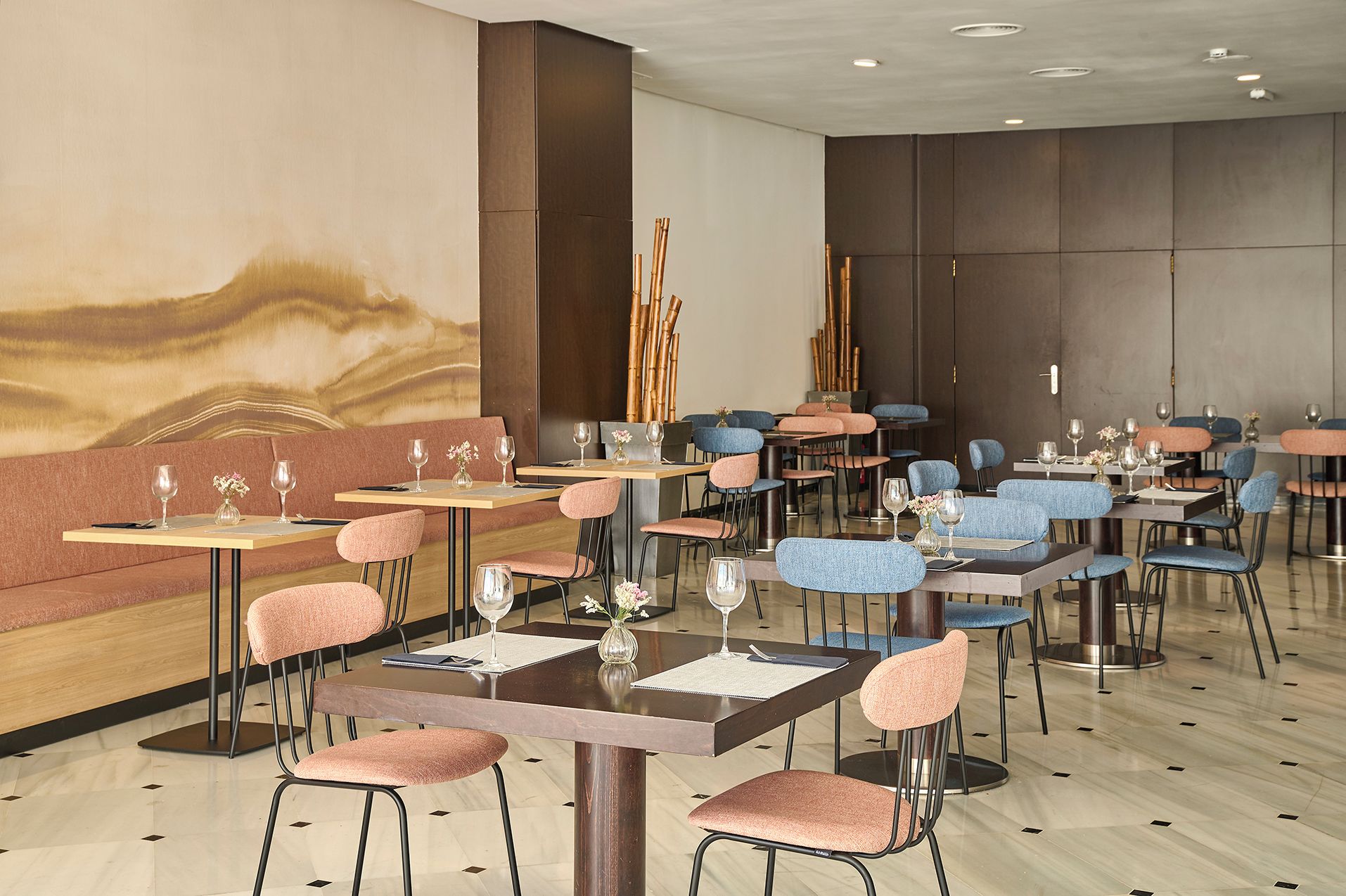 A restaurant with multiple tables and chairs, some with pink and blue cushions, and a large abstract mural on the wall.