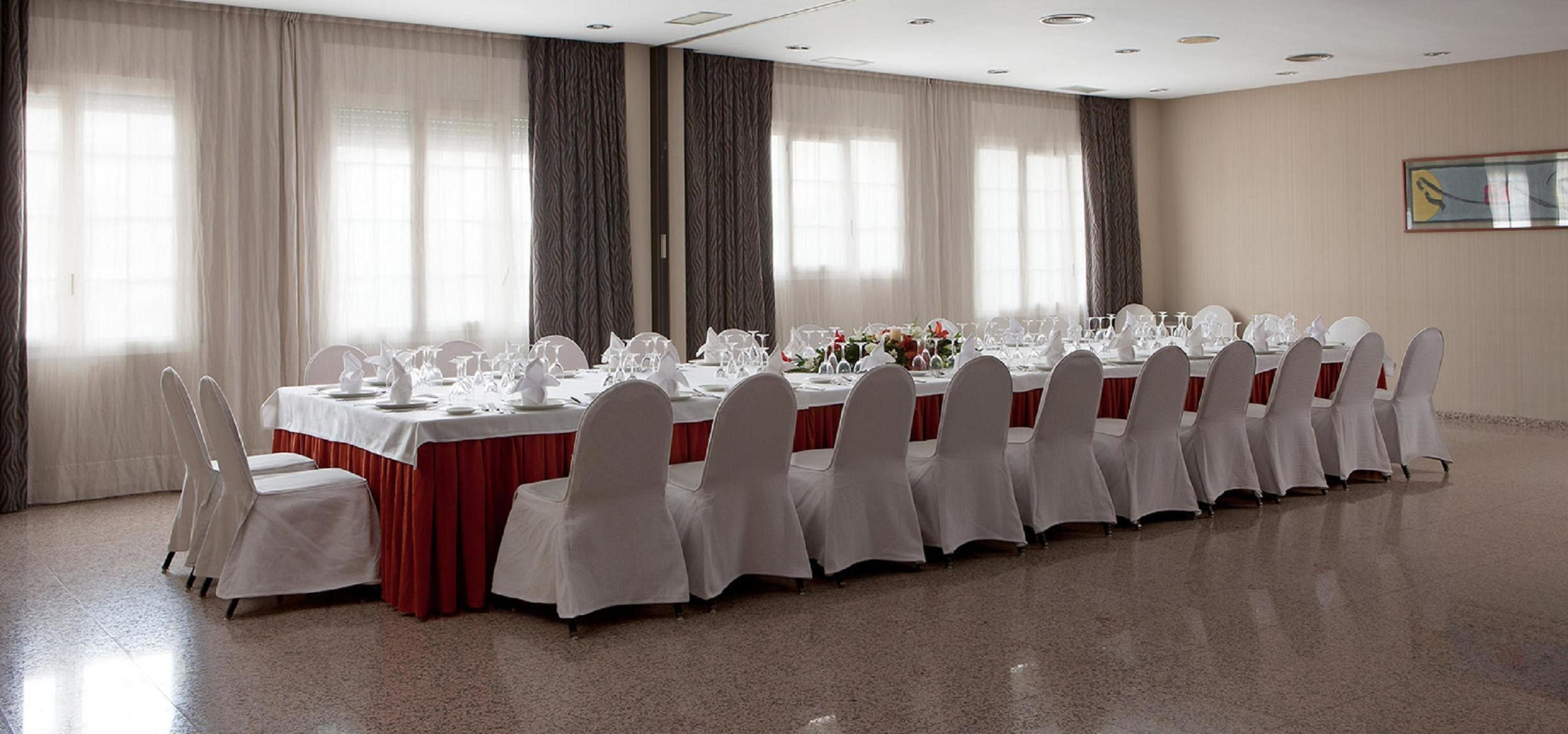 A long banquet table with white tablecloths and matching chairs, set up in a spacious room with large windows and curtains.