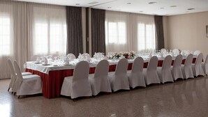 Ein langer Festsaal-Tisch mit weißen Tischdecken und passenden Stühlen, platziert in einem geräumigen Raum mit großen Fenstern und Vorhängen.