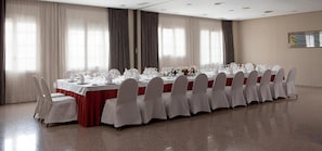 Une longue table de banquet, recouverte de nappes blanches et agrémentée de chaises assorties, est installée dans une pièce spacieuse dotée de grandes fenêtres et de rideaux.