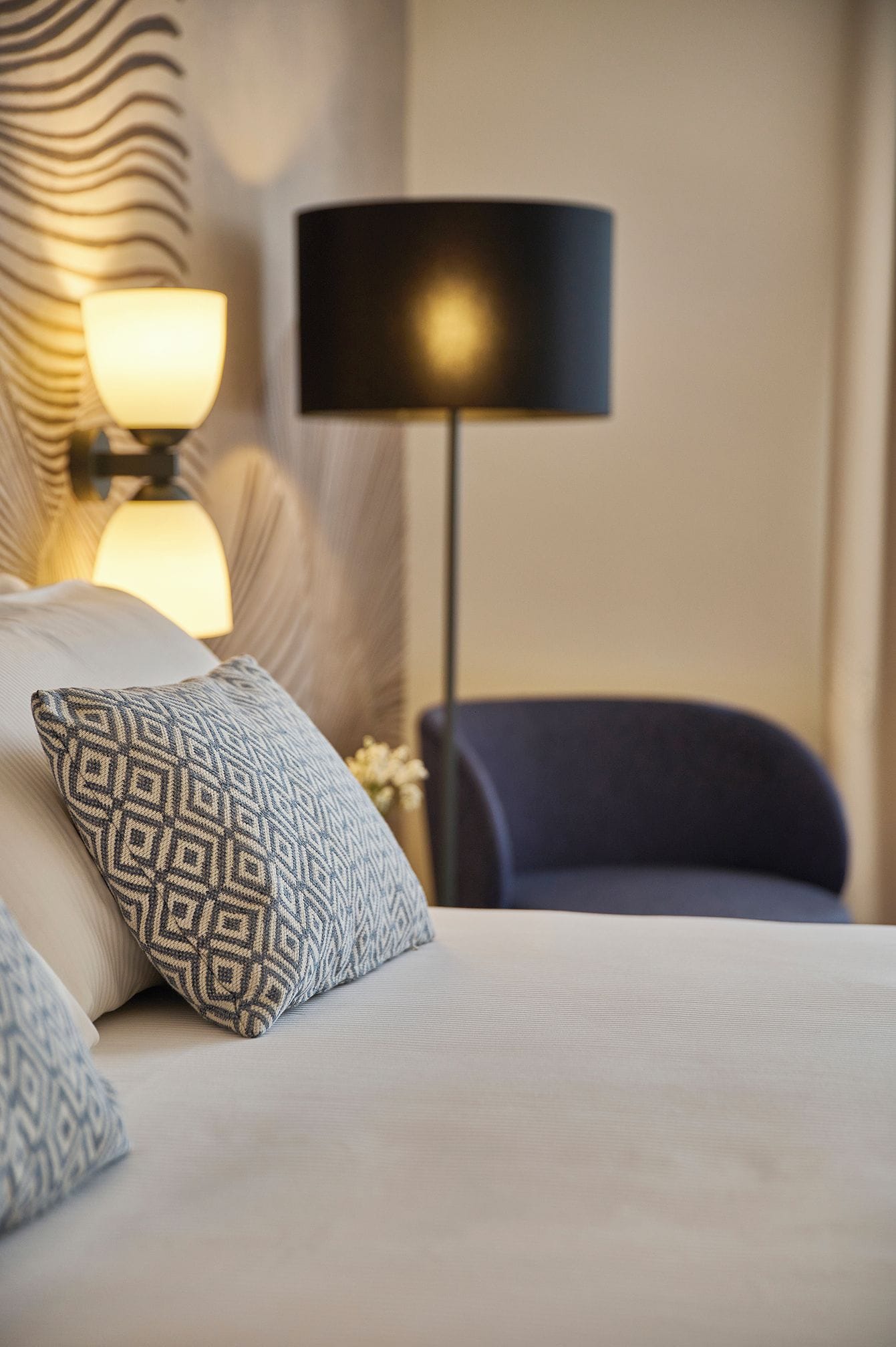 Een net opgemaakt bed met gestreepte kussens, een bedlampje en een stoel in een hotelkamer.