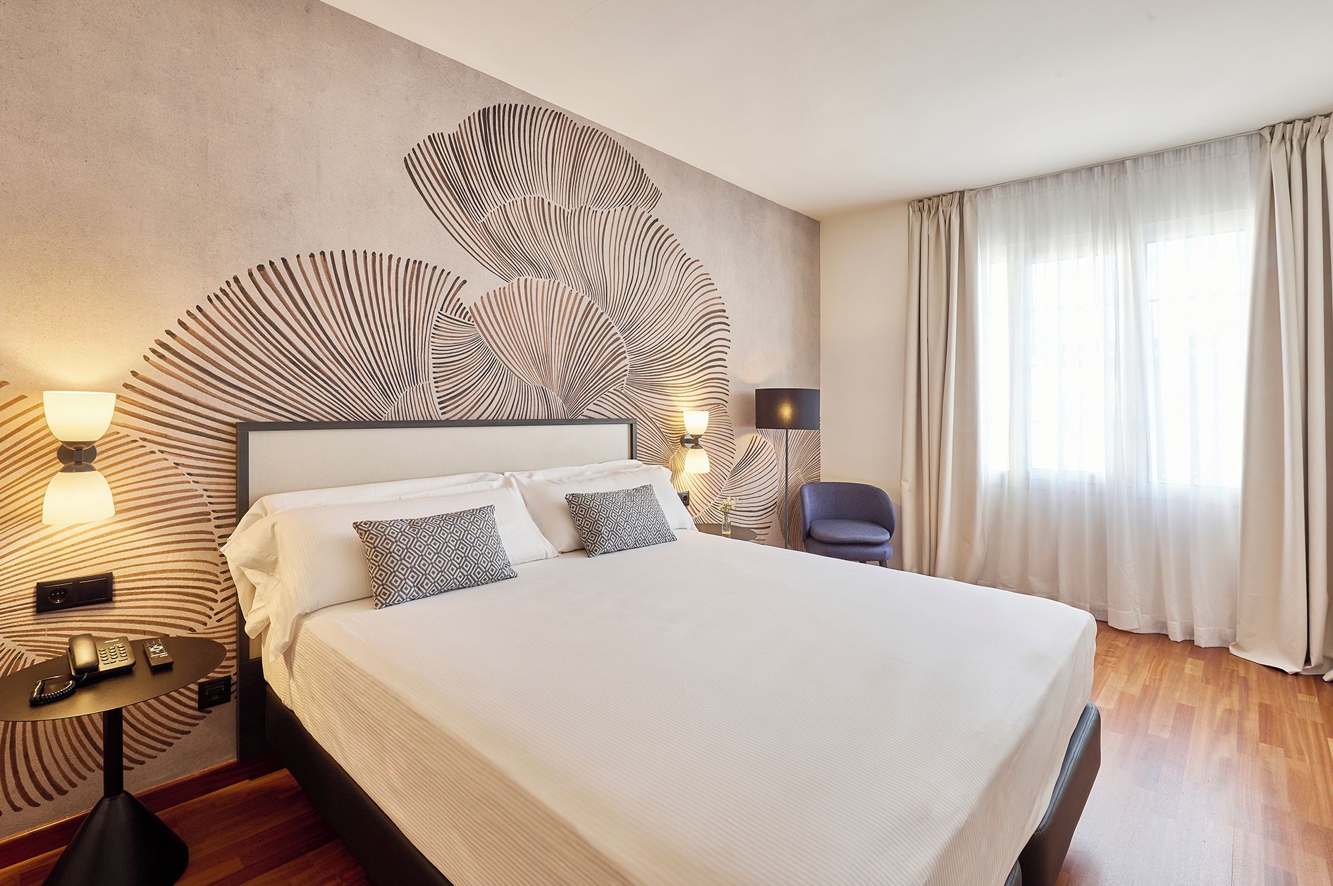Habitación de hotel con una cama grande, una mesita de noche con lámpara, una silla y una pared con diseño de hojas.