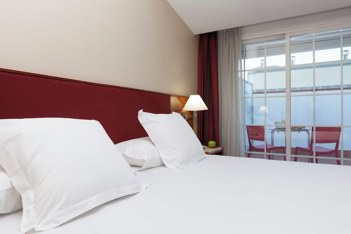 Habitación de hotel con cama, mesita de noche con lámpara, mesa con sillas y vista al mar a través de amplios ventanales.