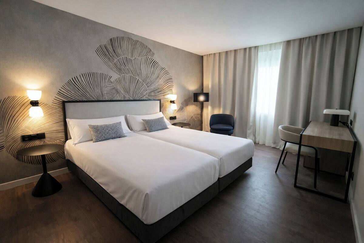 Cama doble con cabecera moderna, lámparas de noche, escritorio y una silla en una habitación de hotel.