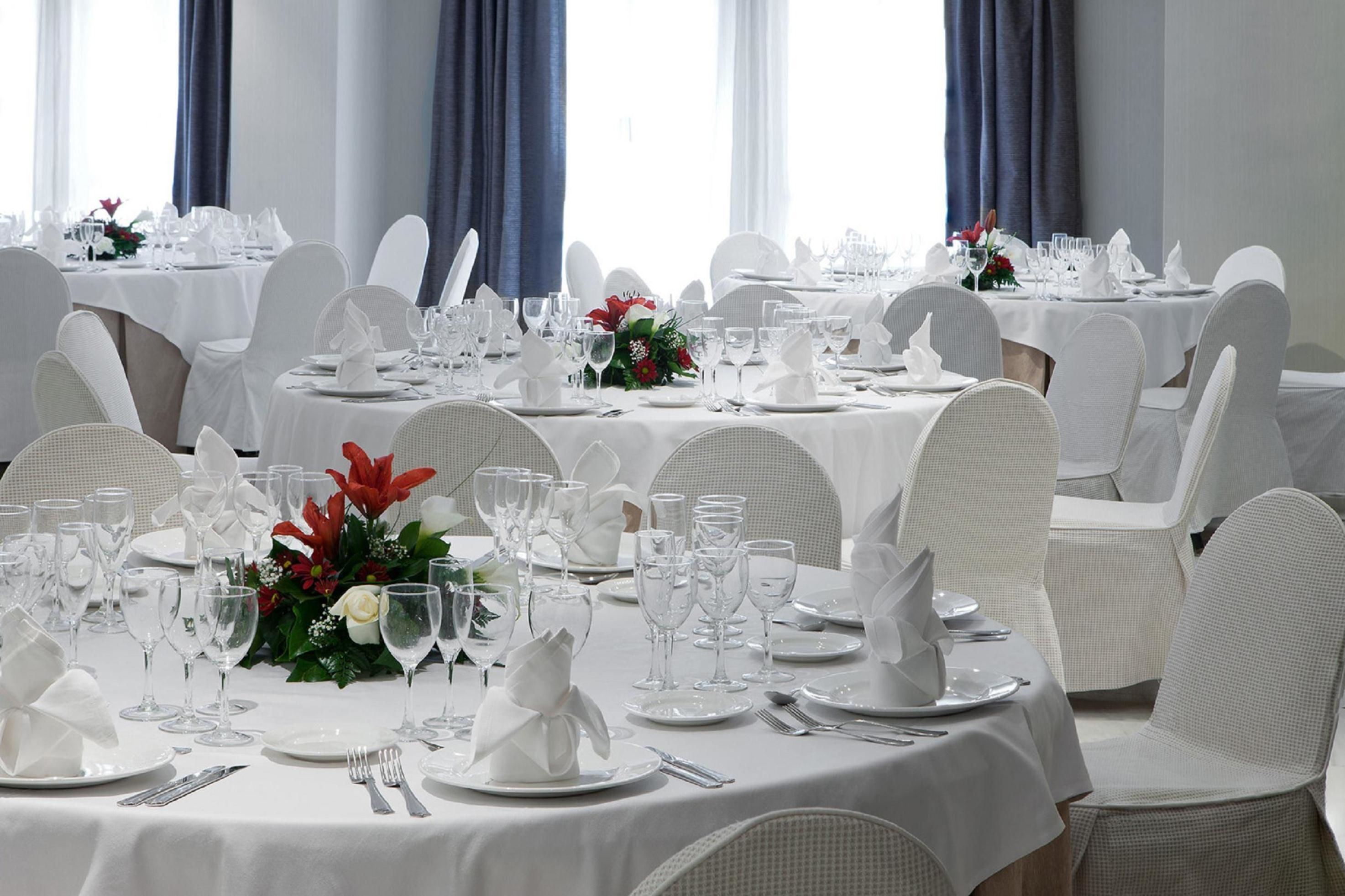 Un salón de banquetes con varias mesas redondas dispuestas para comer, cada una con manteles blancos, sillas blancas y una presentación impecable con platos, vasos y cubiertos.