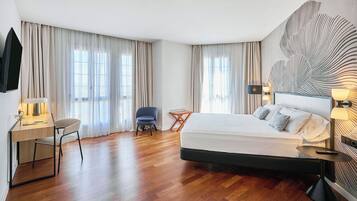 Quarto de hotel com cama, escrivaninha, cadeira, mesa pequena e uma janela grande com cortinas.