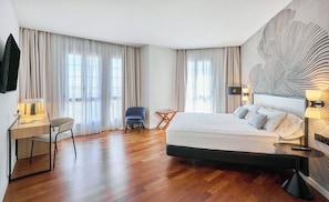Een hotelkamer met een bed, een bureau, een stoel, een bijzettafeltje en een groot raam met gordijnen.