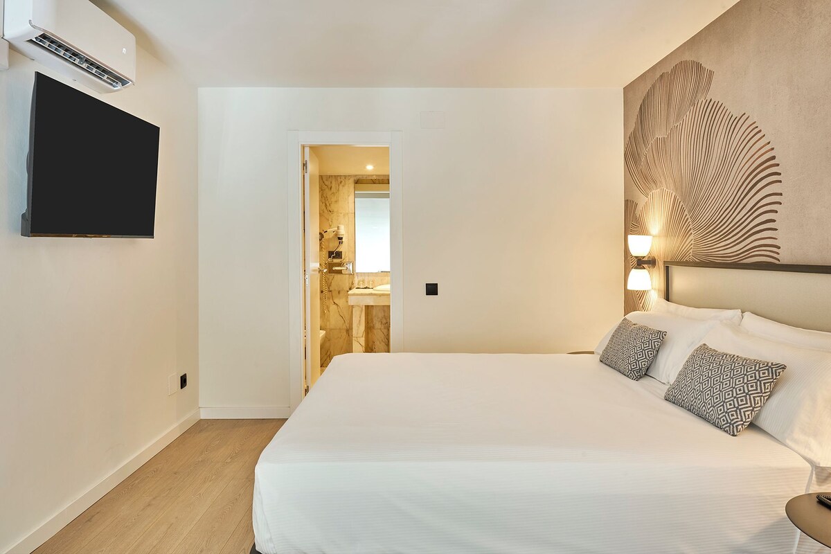 Een hotelkamer met een bed, een wandtelevisie, een badkamer door een geopende deur zichtbaar, en een wandlamp.
