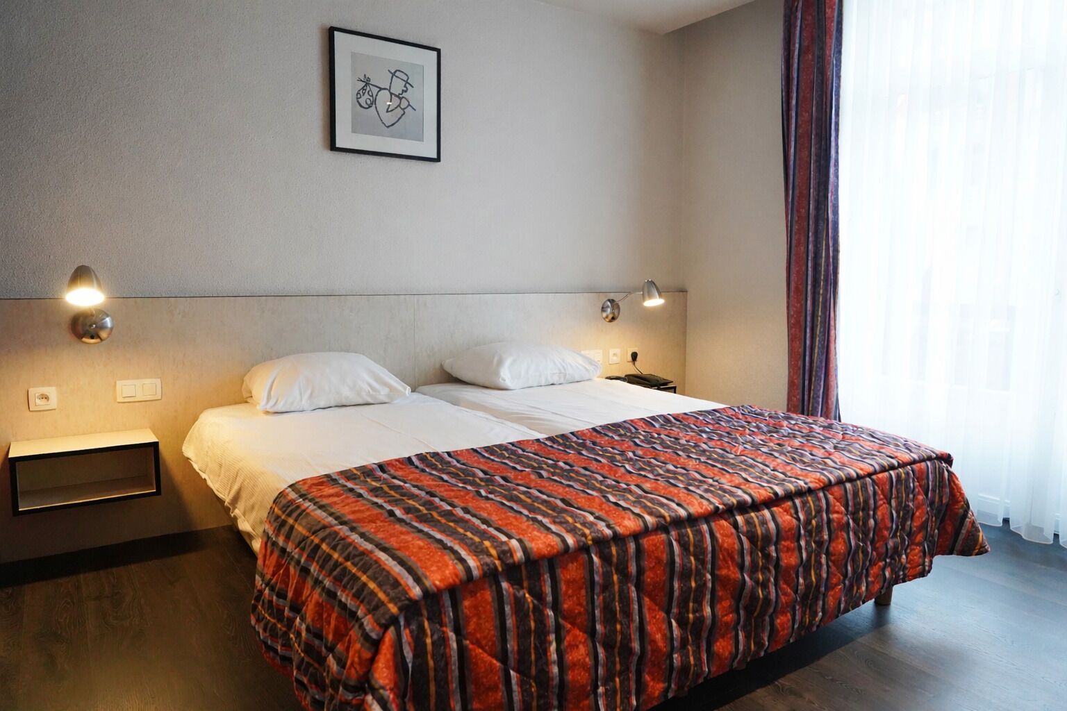 Habitació d'hotel amb un llit, dues tavernes de nit, una estantalla, un quadre al mur i una finestra amb cortines.