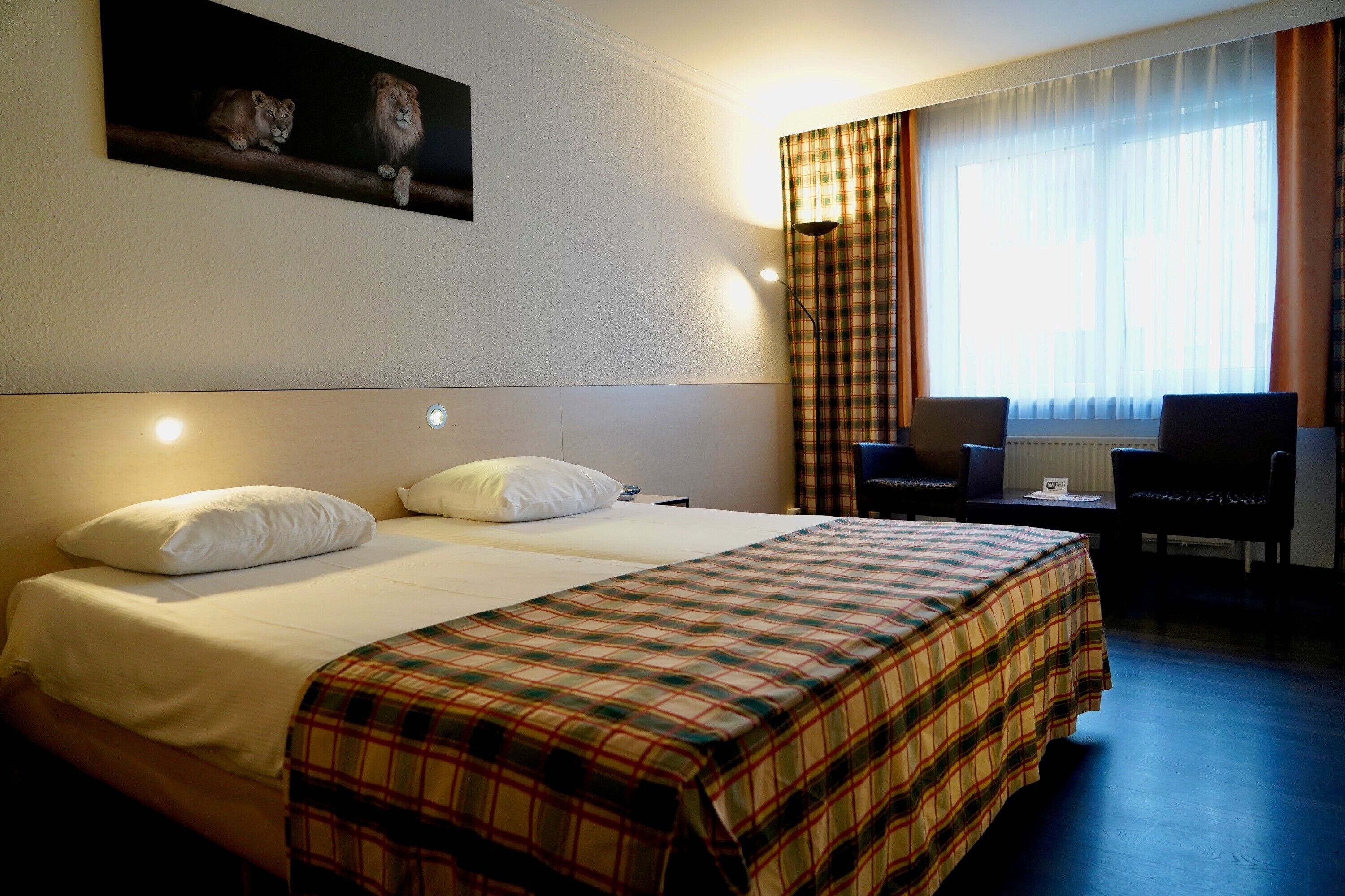 Une chambre d’hôtel avec un grand lit, deux oreillers, une couverture à carreaux, un bureau avec deux chaises et une fenêtre avec des rideaux.