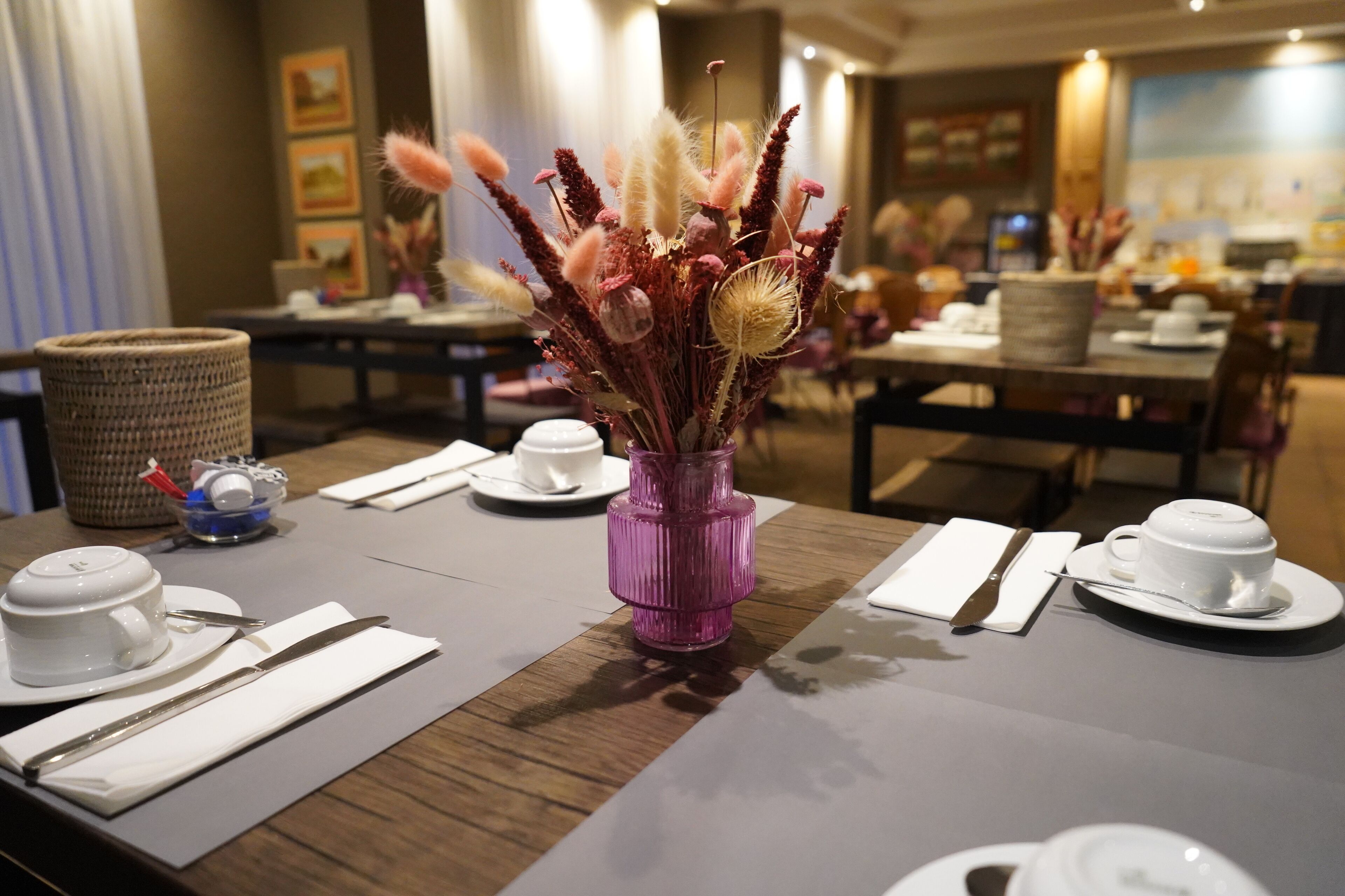 Une table à manger dressée avec des tasses, des soucoupes et des couverts blancs, un vase violet contenant des fleurs séchées et un panier tressé.