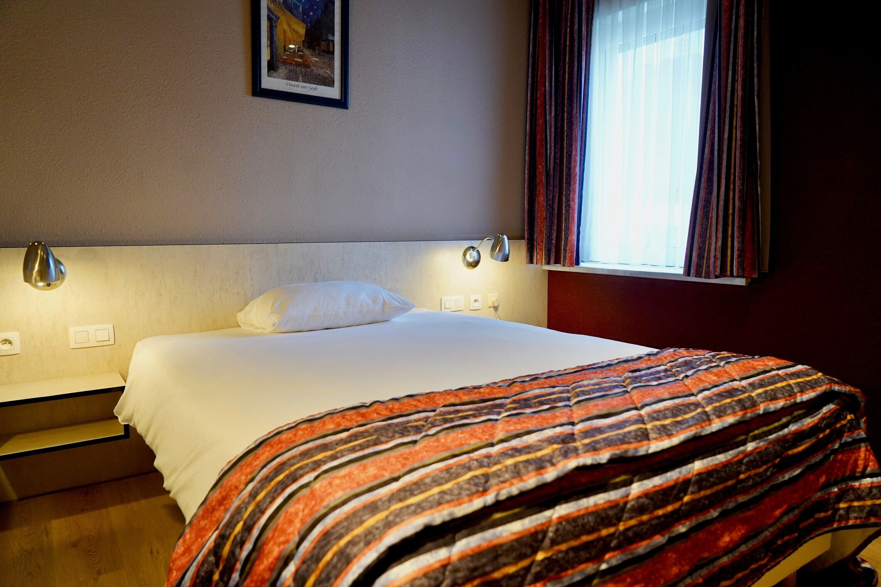 Une chambre d’hôtel avec un lit, une table de chevet, une lampe, une fenêtre avec des rideaux et un tableau au mur.