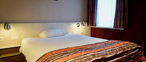 A hotel room with a bed, a nightstand, a lamp, a window with curtains, and a painting on the wall.