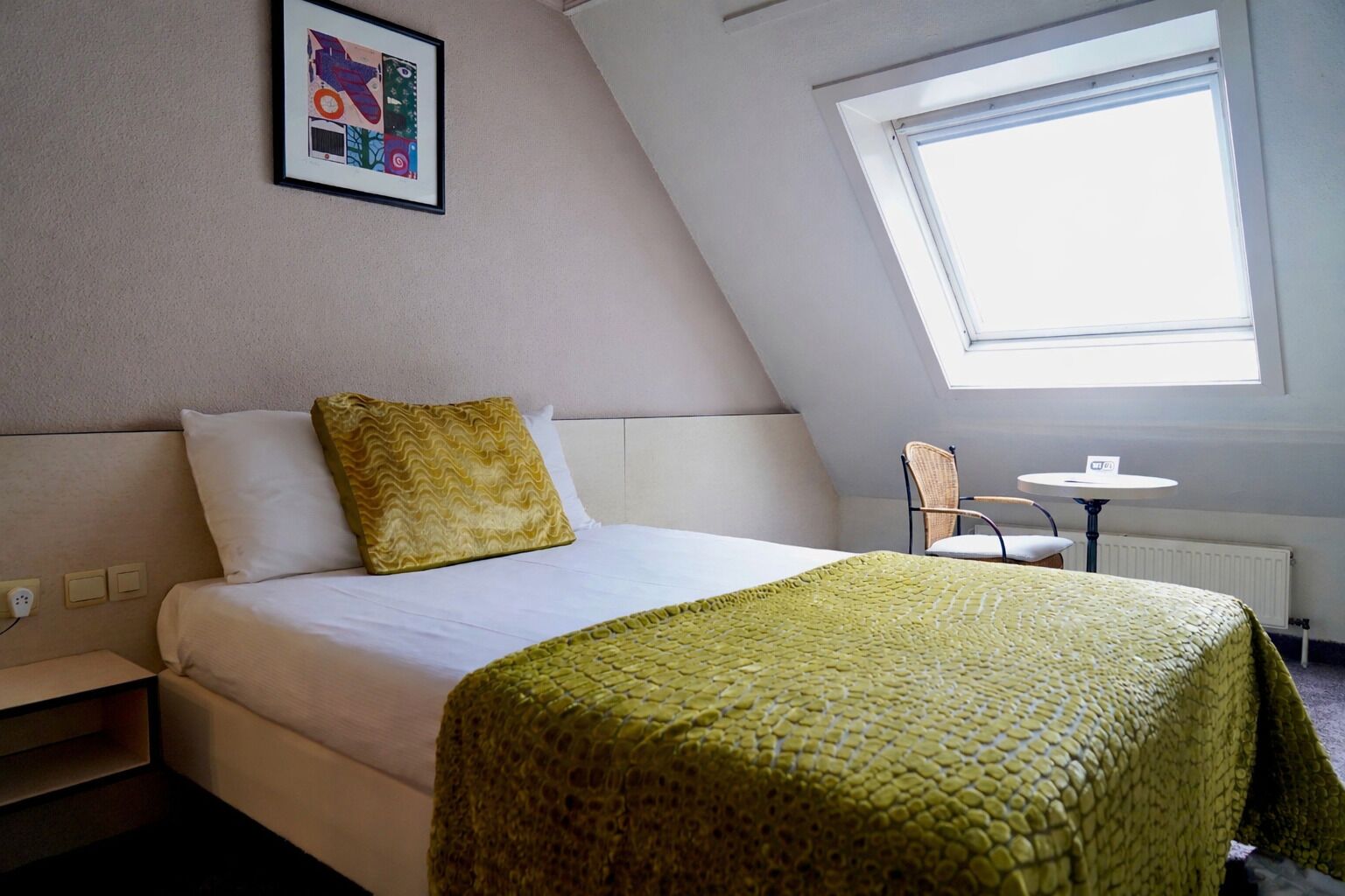 Habitación de hotel con cama, manta verde con textura, almohada blanca, mesita pequeña y una silla. Se observa un cuadro en la pared y una ventana que proporciona luz natural.
