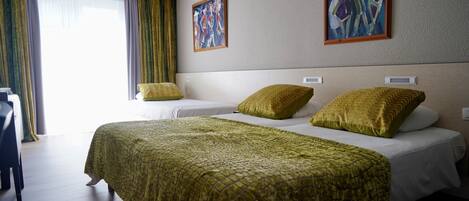 Habitación de hotel con una cama grande, dos camas más pequeñas, un escritorio y una silla. Hay dos cuadros en la pared y una ventana con cortinas.