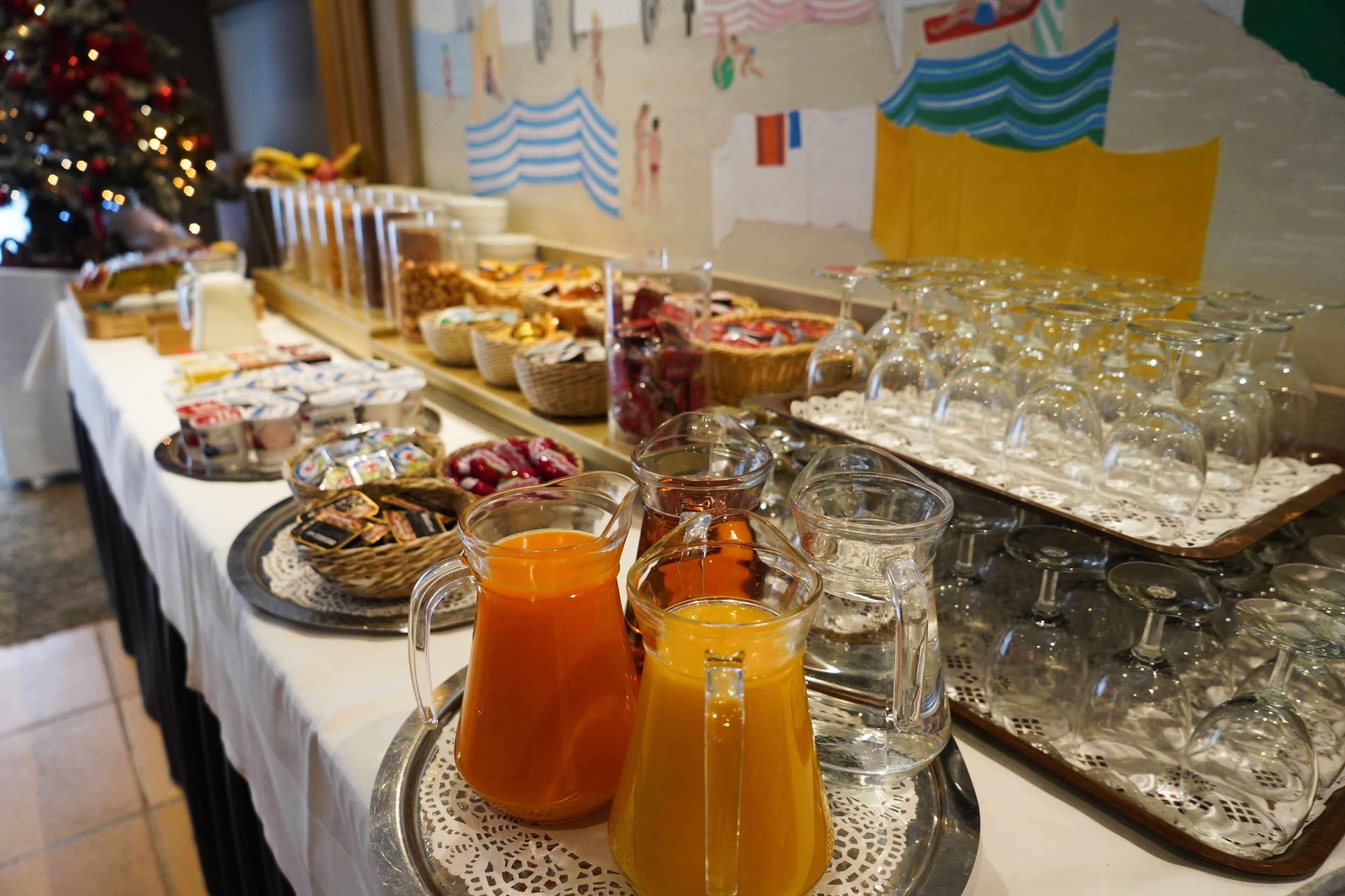 Mesa de buffet com diversos alimentos e bebidas, incluindo uma árvore de Natal ao fundo.