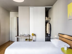 Une chambre d’hôtel avec un lit, un bureau, une chaise, une armoire et une petite table avec une tasse.