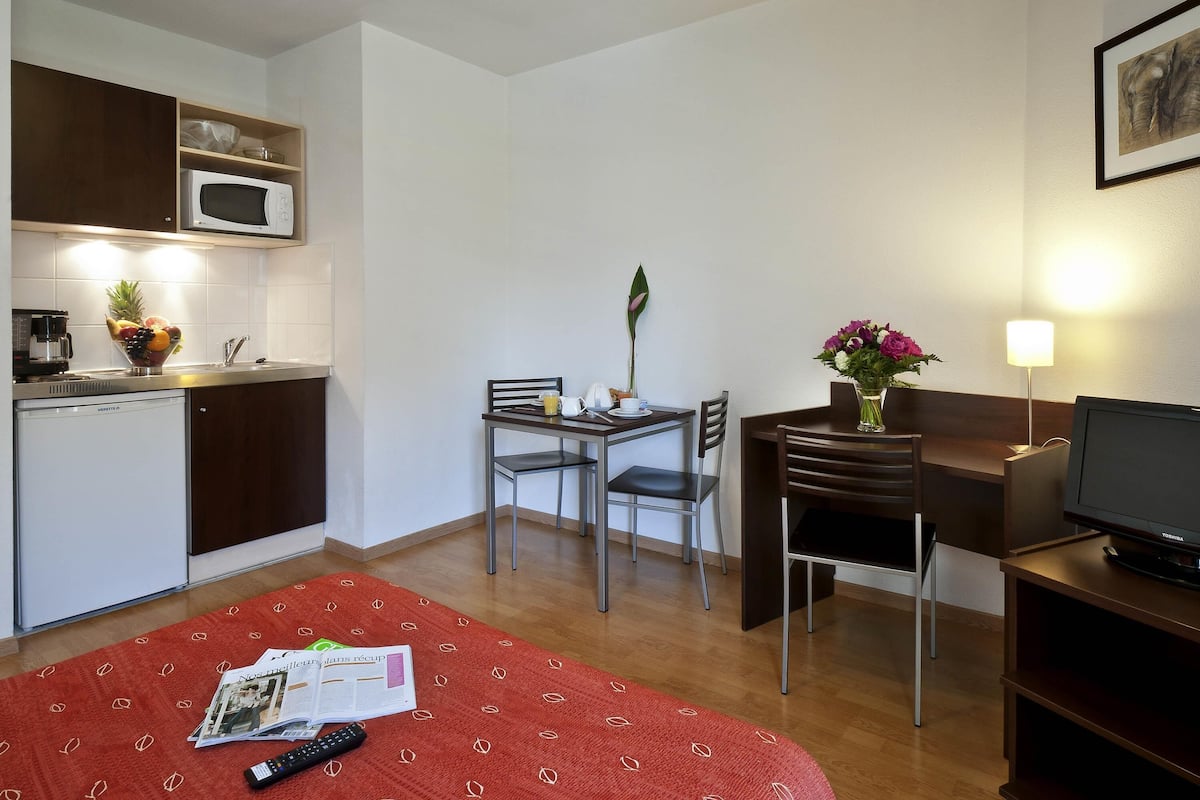 Une chambre d’hôtel compacte comprenant un coin cuisine, un espace repas, une télévision et un lit agrémenté d’un tapis à motifs rouges.