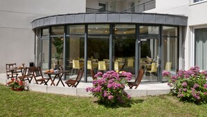 Ein modernes Gebäude mit einer gewölbten Glasfassade, einem Außenbereich mit Tischen und Stühlen sowie einem gepflegten Garten mit rosa Blüten.
