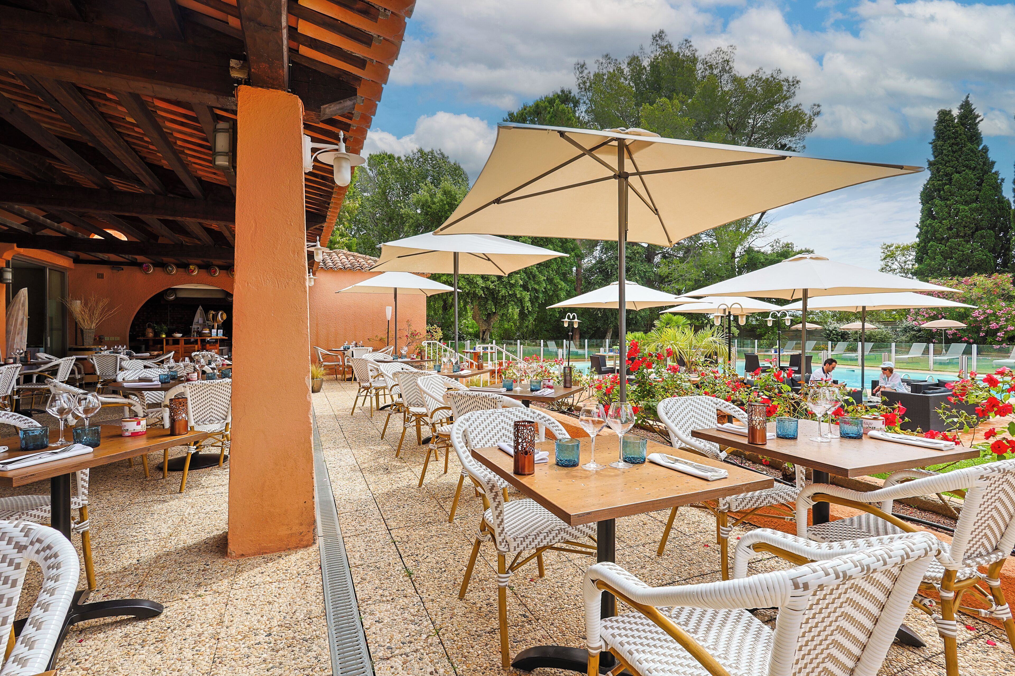 An outdoor patio with wicker chairs and tables, large umbrellas, and a pool area in the background.