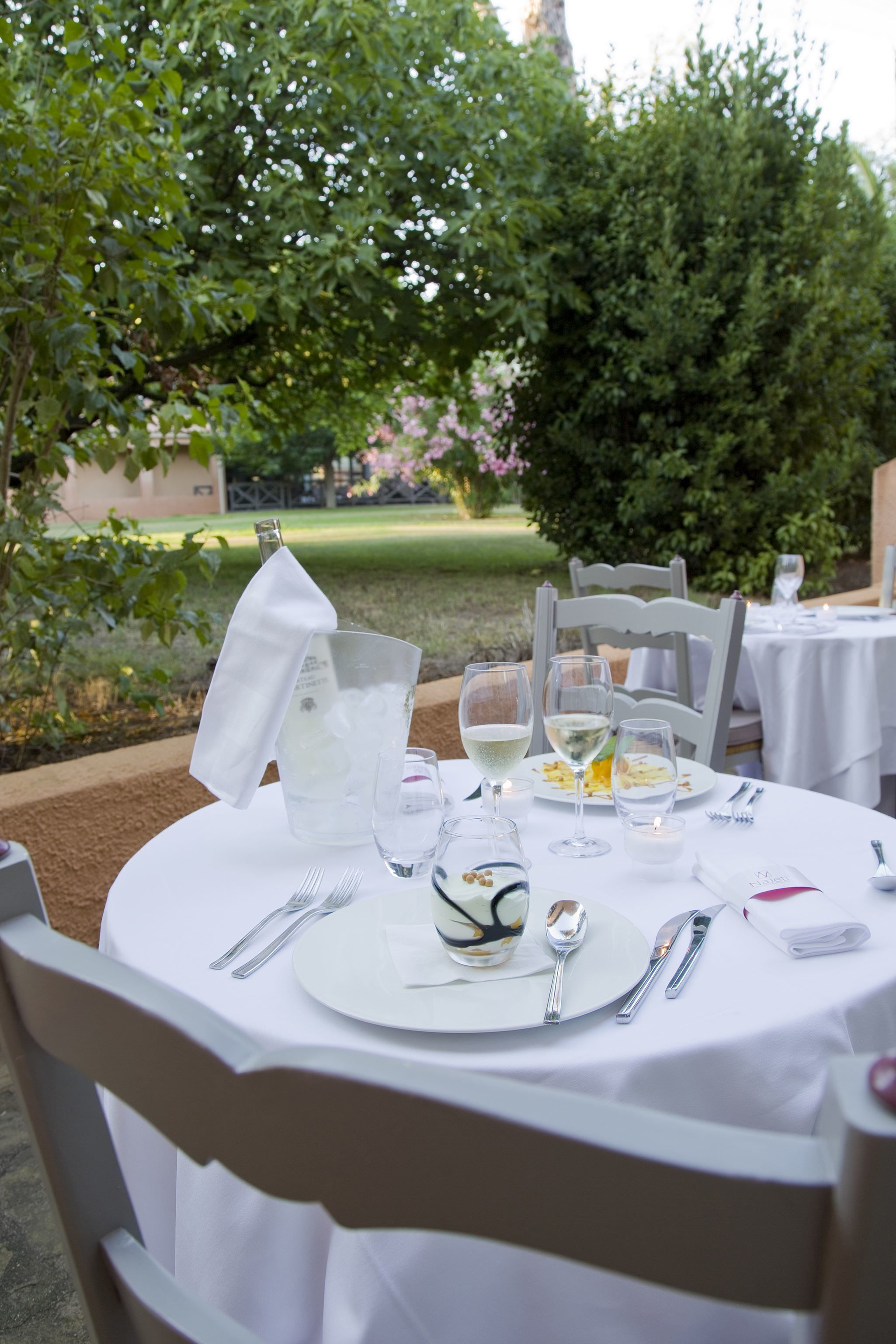 Un área de comedor al aire libre con manteles blancos, una botella de champán y una copa de vino blanco.