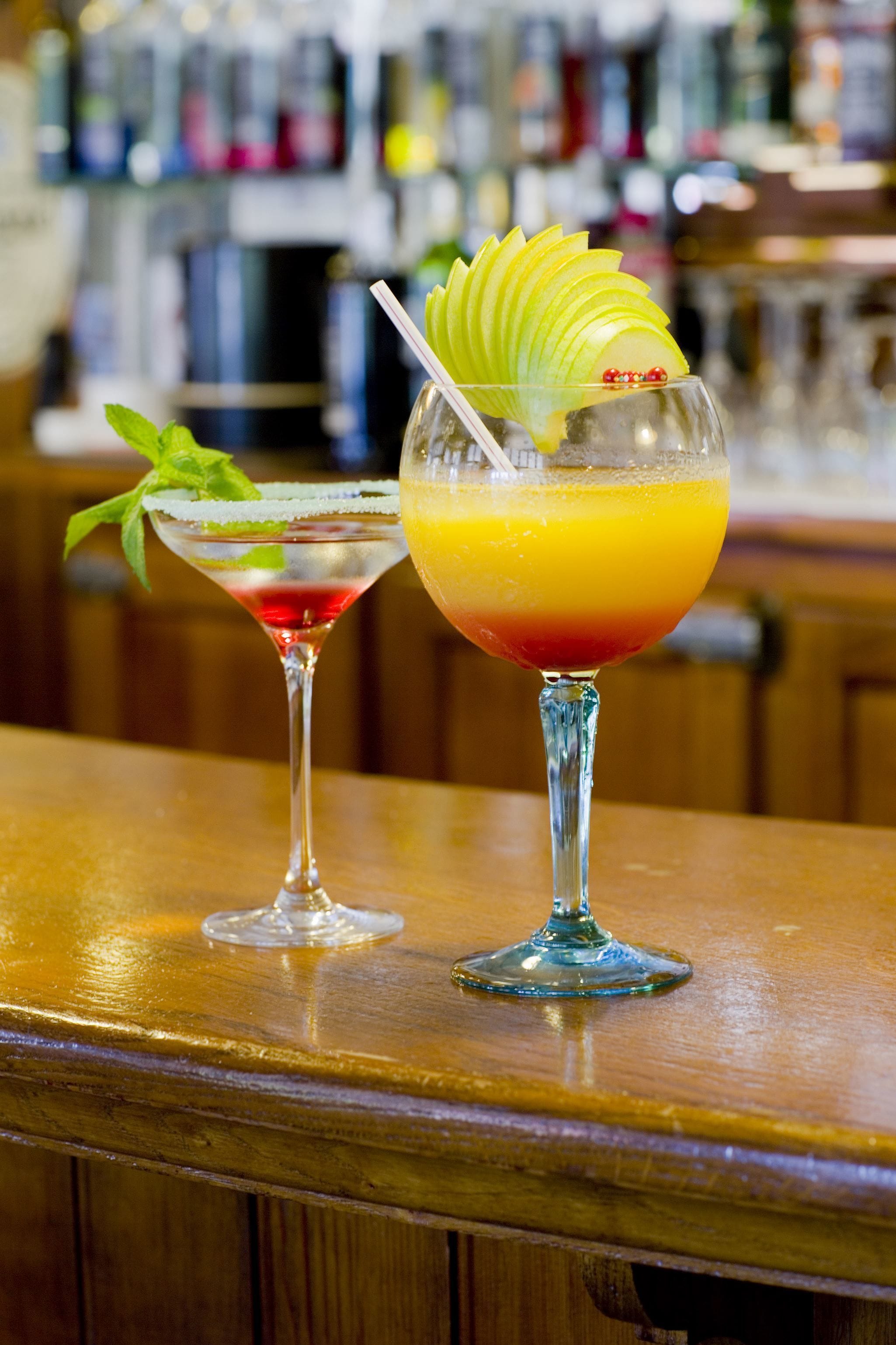 Two cocktails on a bar counter with a blurred background of bottles.
